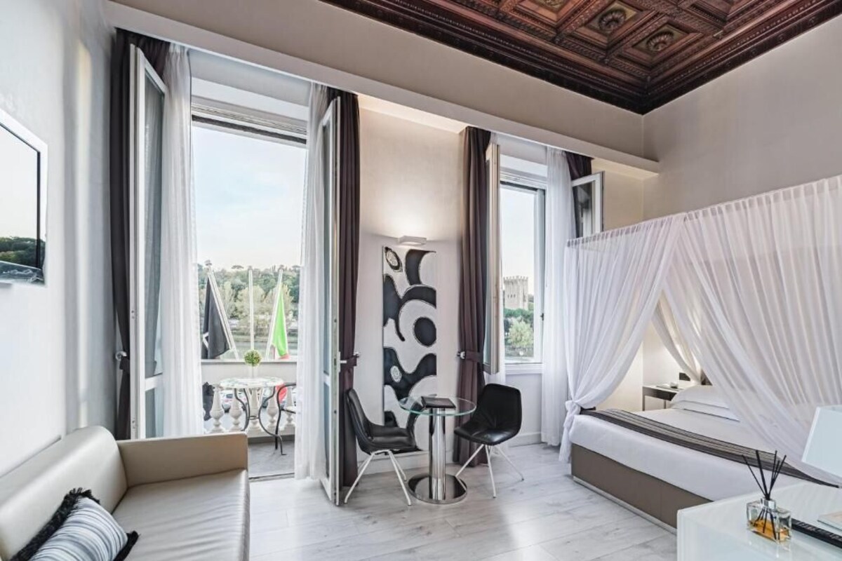 A modern hotel room with a large bed, a sitting area with a table and chairs, and a balcony with a view of the city.