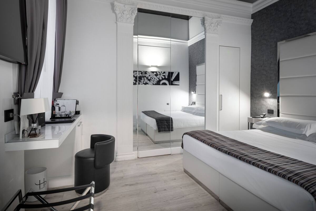 A modern hotel room with a large bed, a desk with a lamp, a chair, and a mirror.