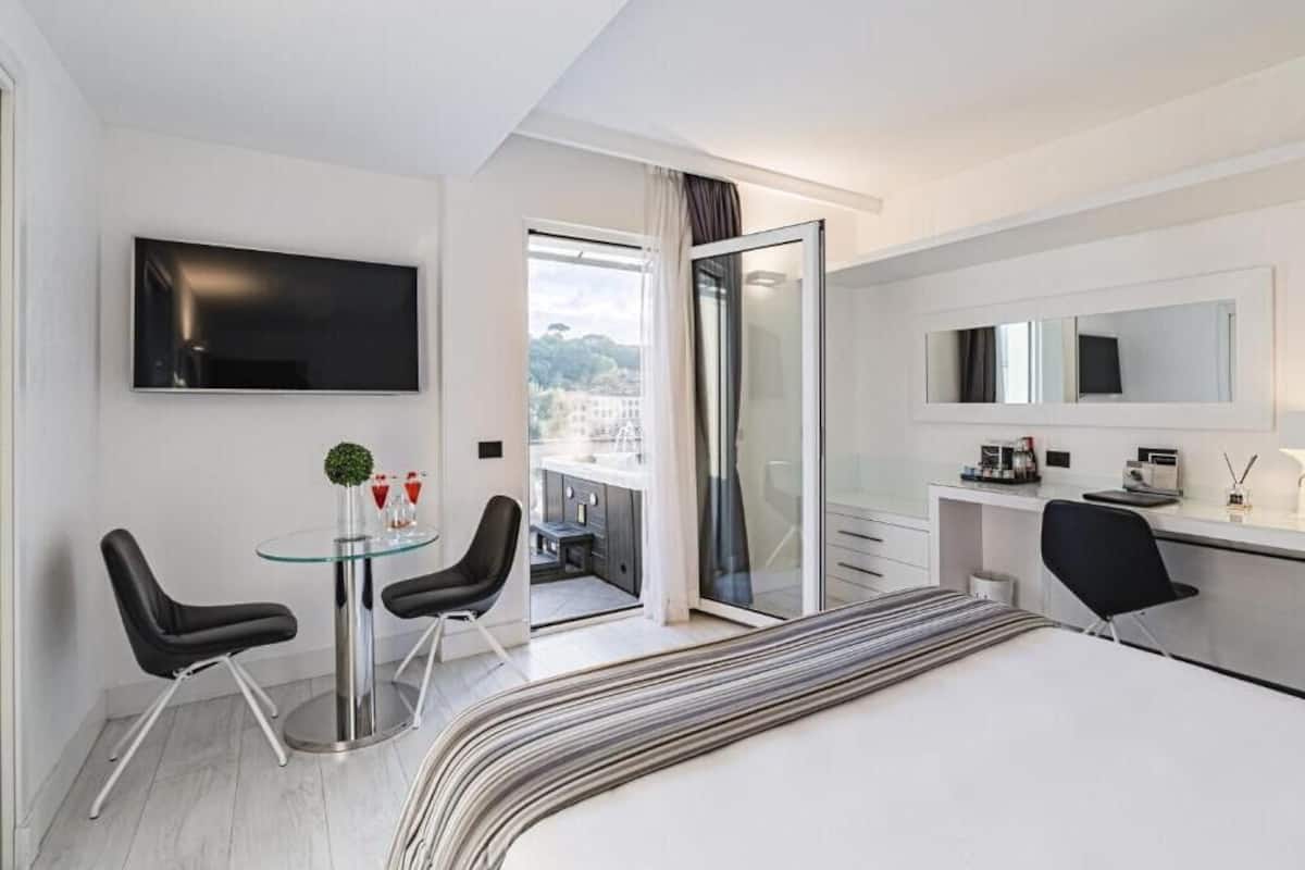 A modern hotel room with a large bed, a glass dining table with chairs, a TV mounted on the wall, a sliding glass door leading to a balcony, and a small kitchenette.
