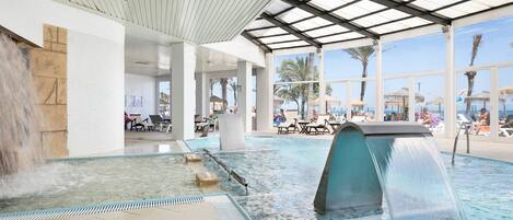 En indendørs swimmingpool med et vandfald, liggestole og udsigt til palmer udenfor.