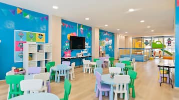 Een vrolijk ingerichte kinderspeelruimte met tafels en stoelen, educatieve posters en een televisie.