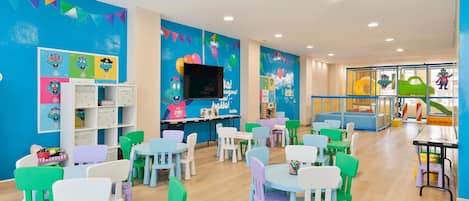 Une salle de jeux pour enfants aux couleurs vives, avec des tables et des chaises, des affiches éducatives et un téléviseur.
