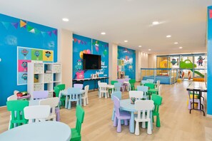 Un salón de juegos infantil, lleno de colores, con mesas, sillas, carteles educativos y un televisor.