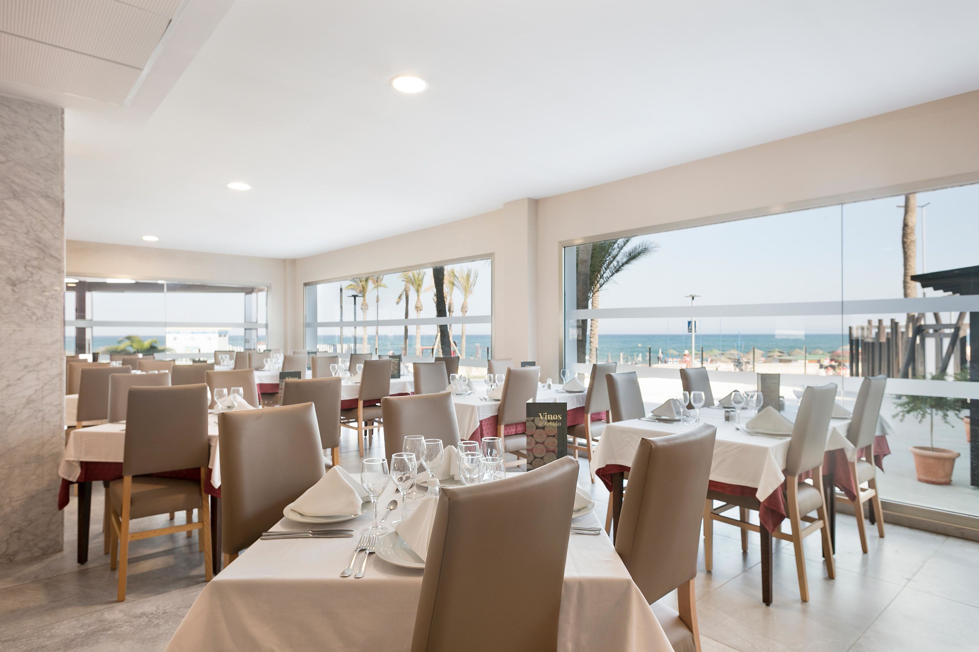 A dining area with tables set for service, large windows offering a view of the beach, and a calm sea in the background.