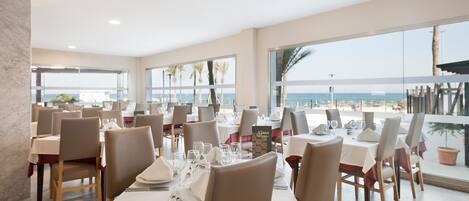 A dining area with tables set for service, large windows offering a view of the beach, and a calm sea in the background.