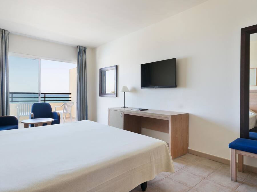 Habitación de hotel con cama, escritorio, silla, televisión y balcón con cortinas.