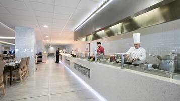 A modern kitchen area with chefs preparing food, a buffet setup, and a dining area with tables and chairs.