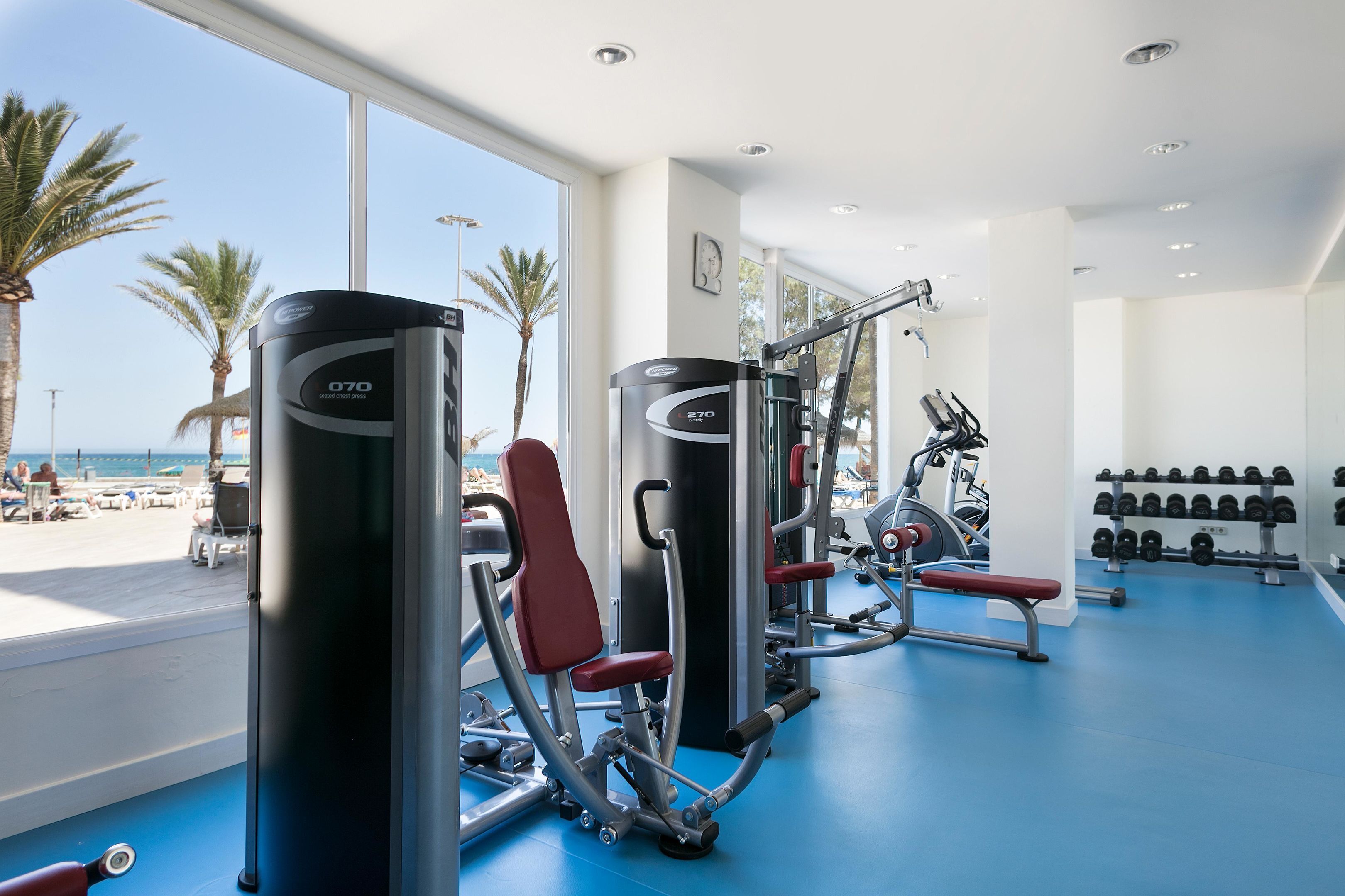 Un gimnasio con equipo de ejercicio, suelo azul y vistas a la playa a través de amplias ventanas.