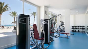 Un gimnasio con equipo de ejercicio, suelo azul y vistas a la playa a través de amplias ventanas.
