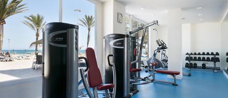 A gym with exercise equipment, a blue floor, and a view of the beach through large windows.