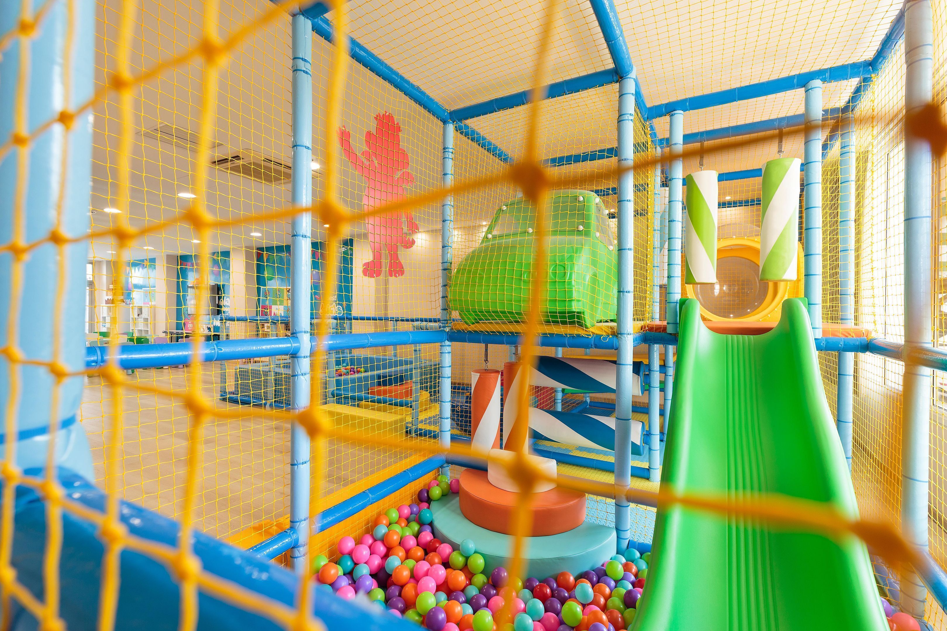 Een speelruimte met een groene glijbaan, kleurrijke ballen en een omheining met net.