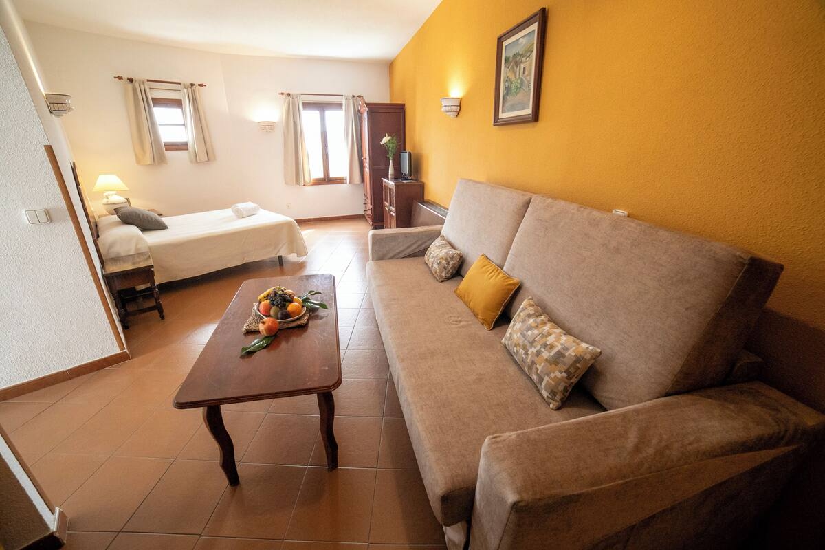 A living room with a sofa, a small table with fruits, a bed, and a TV.