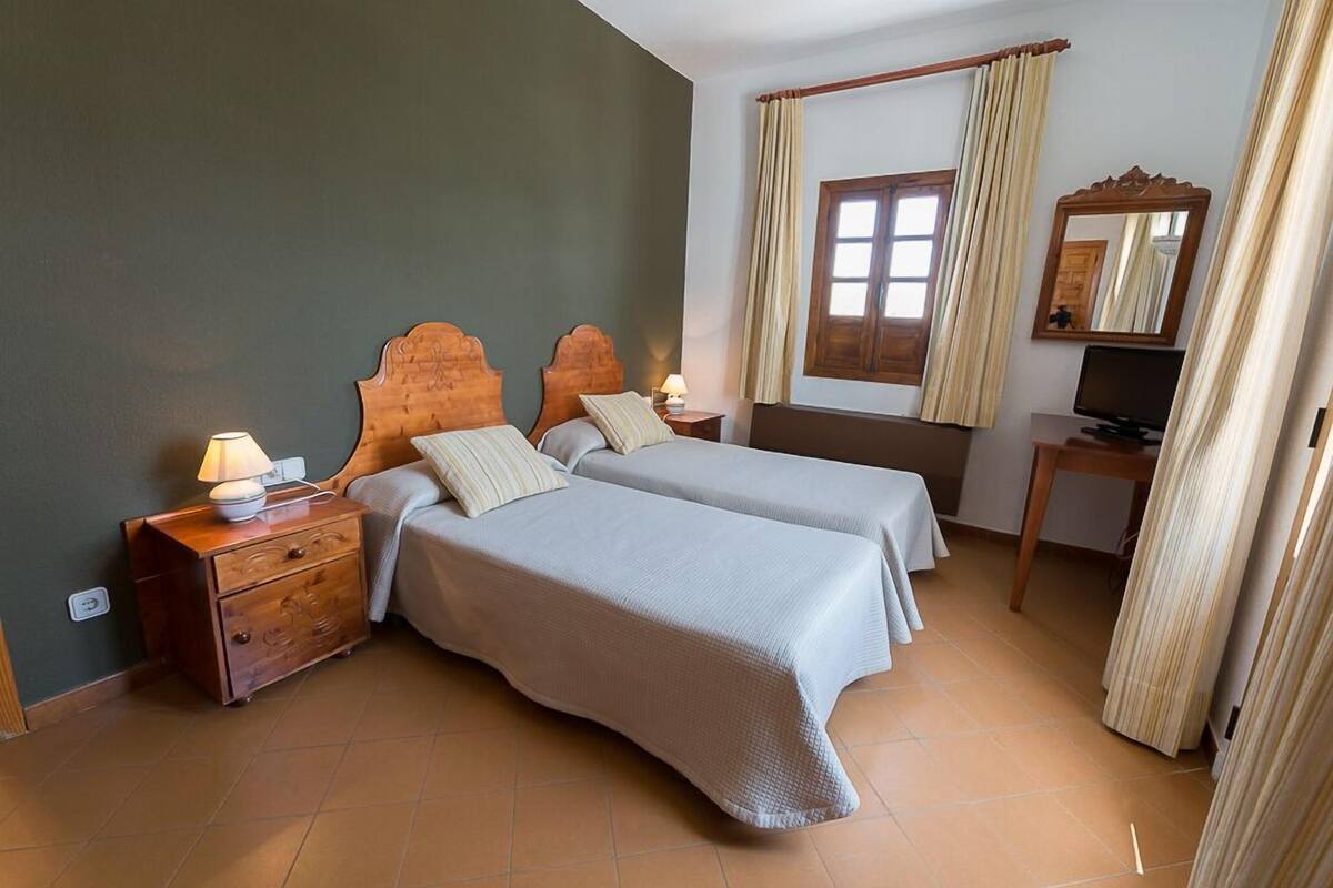 A bedroom with two beds, a wooden nightstand with a lamp, a desk with a computer, and a window with curtains.