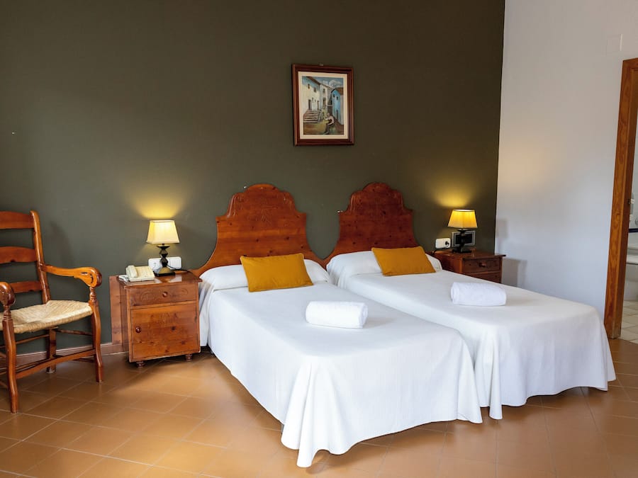 A hotel room with two beds, a wooden chair, a nightstand with a lamp, and a painting on the wall.