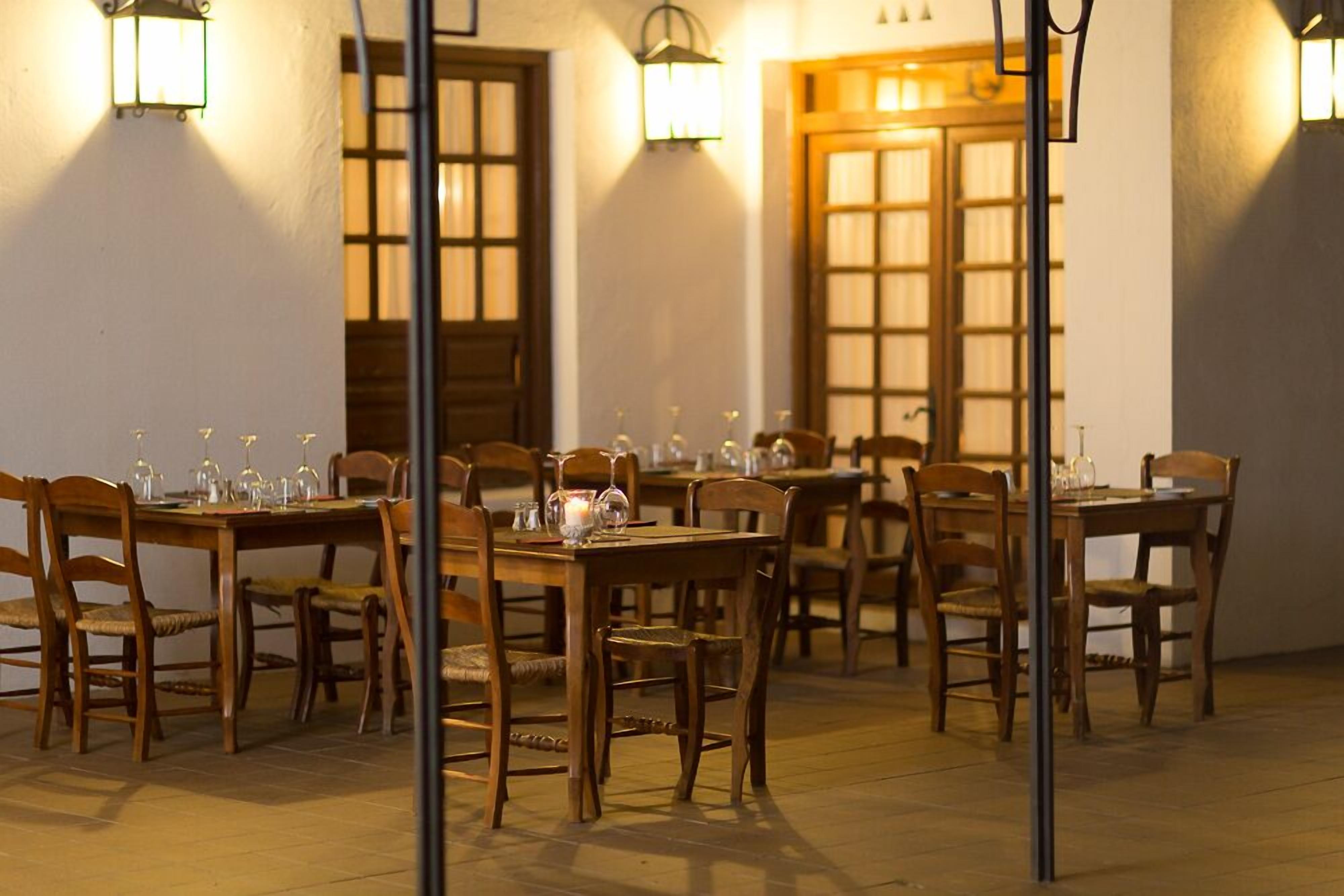 A dining area with wooden tables and chairs, equipped with wine glasses and cutlery, and illuminated by wall-mounted lamps.
