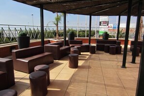 An outdoor seating area with multiple sofas and round tables, a tiled floor, and a view of the cityscape. - Hotel Albufera (Alfafar)