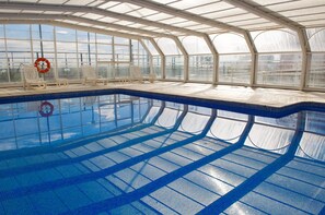Ein überdachter Pool mit großen Glaswänden, Fliesenboden und mehreren Liegestühlen.