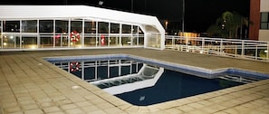 Una piscina con cubierta de madera, rodeada por una barandilla blanca, y un edificio con amplios ventanales al fondo.