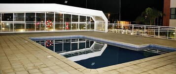 A swimming pool with a wooden deck, surrounded by a white railing, and a building with large windows in the background.