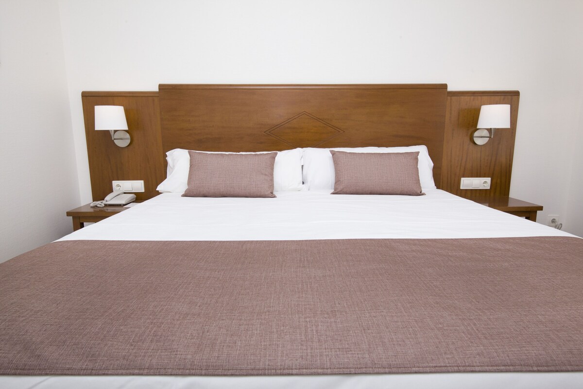 Una cama bien hecha con cabecera de madera, dos lámparas de noche y un teléfono en la mesita.