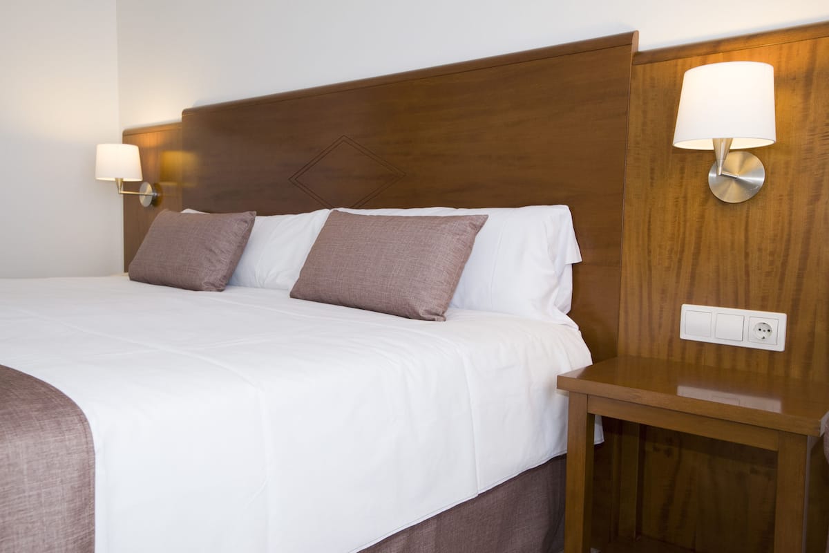 A neatly made bed with white and brown linens, a wooden headboard, and two wall-mounted lamps.