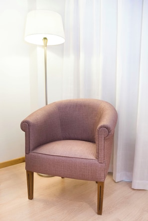 A pink armchair with wooden legs, positioned in front of a floor lamp and white curtains. - Hotel Albufera (Alfafar)