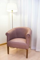 Un sillón rosa con patas de madera, ubicado frente a una lámpara de pie y cortinas blancas.