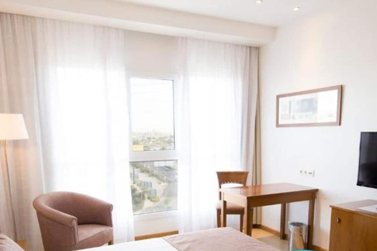 A hotel room with a bed, a chair, a desk, a TV, and a window with curtains.