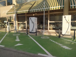 A building with a green parking space marked for electric vehicle charging, a storefront with a striped awning, and a glass facade. - Hotel Albufera (Alfafar)