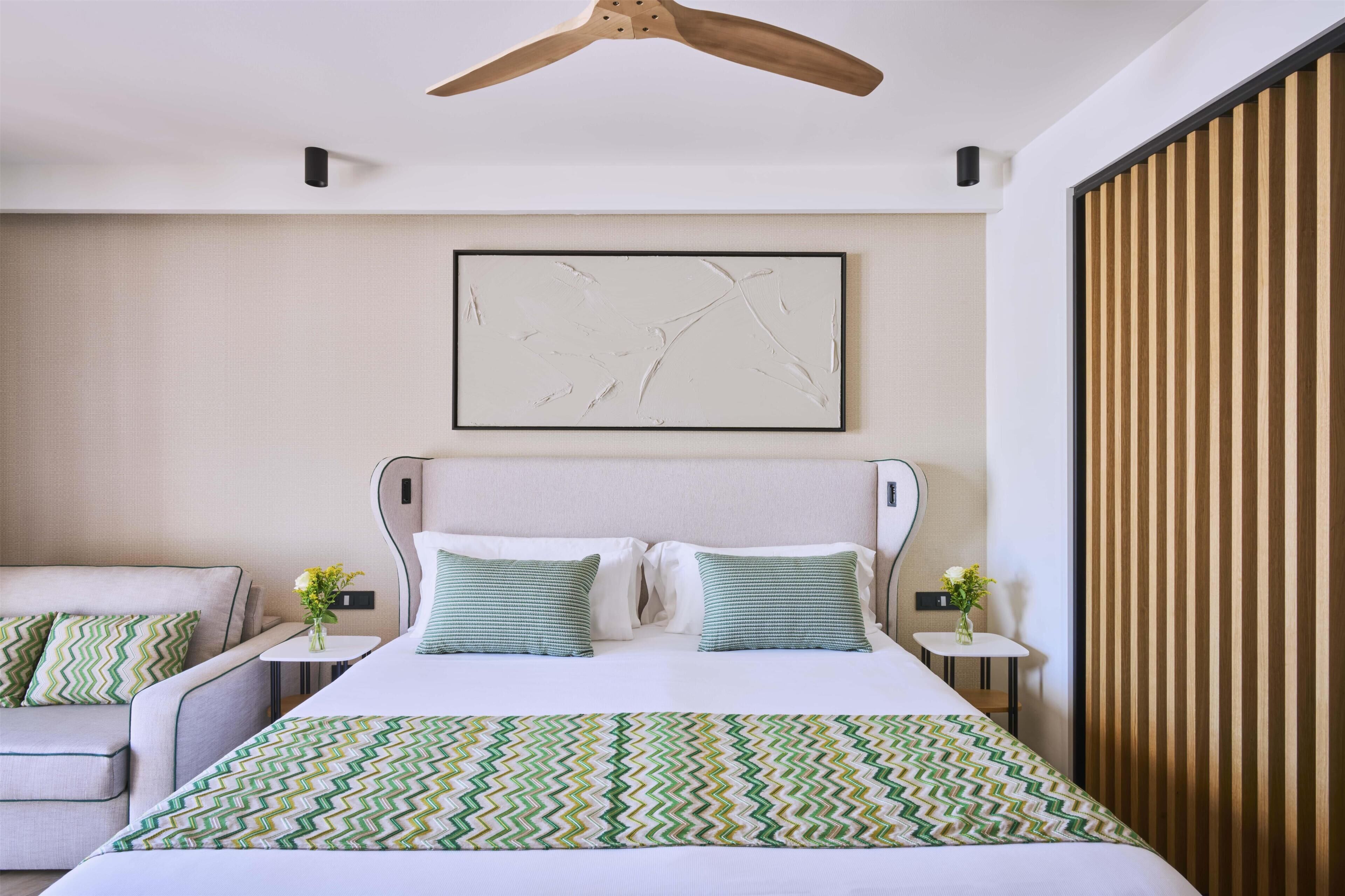 Habitación de hotel con cama, una silla, una mesita y una obra de arte en la pared.