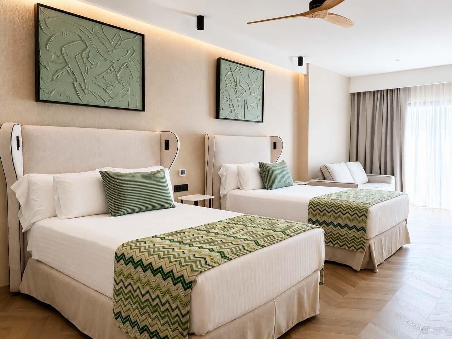 Habitación de hotel con dos camas, ventilador de techo, obras de arte en las paredes y una ventana con cortinas.