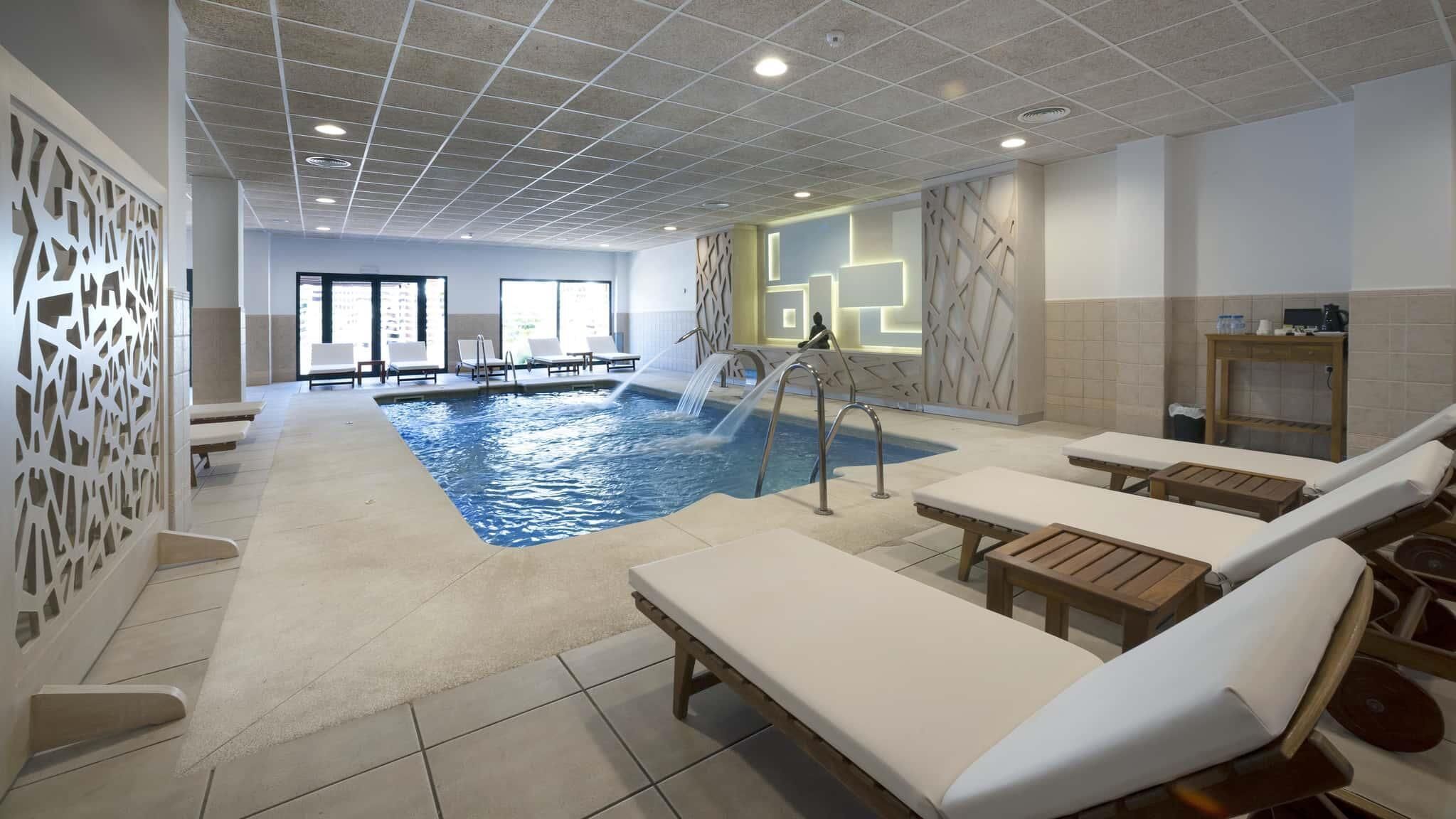 A modern indoor swimming pool with lounge chairs, a water slide, and a reception area.