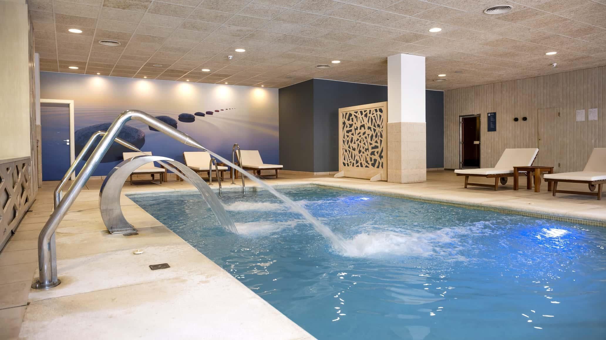 An indoor swimming pool with a metal ladder, lounge chairs, and a decorative wall.