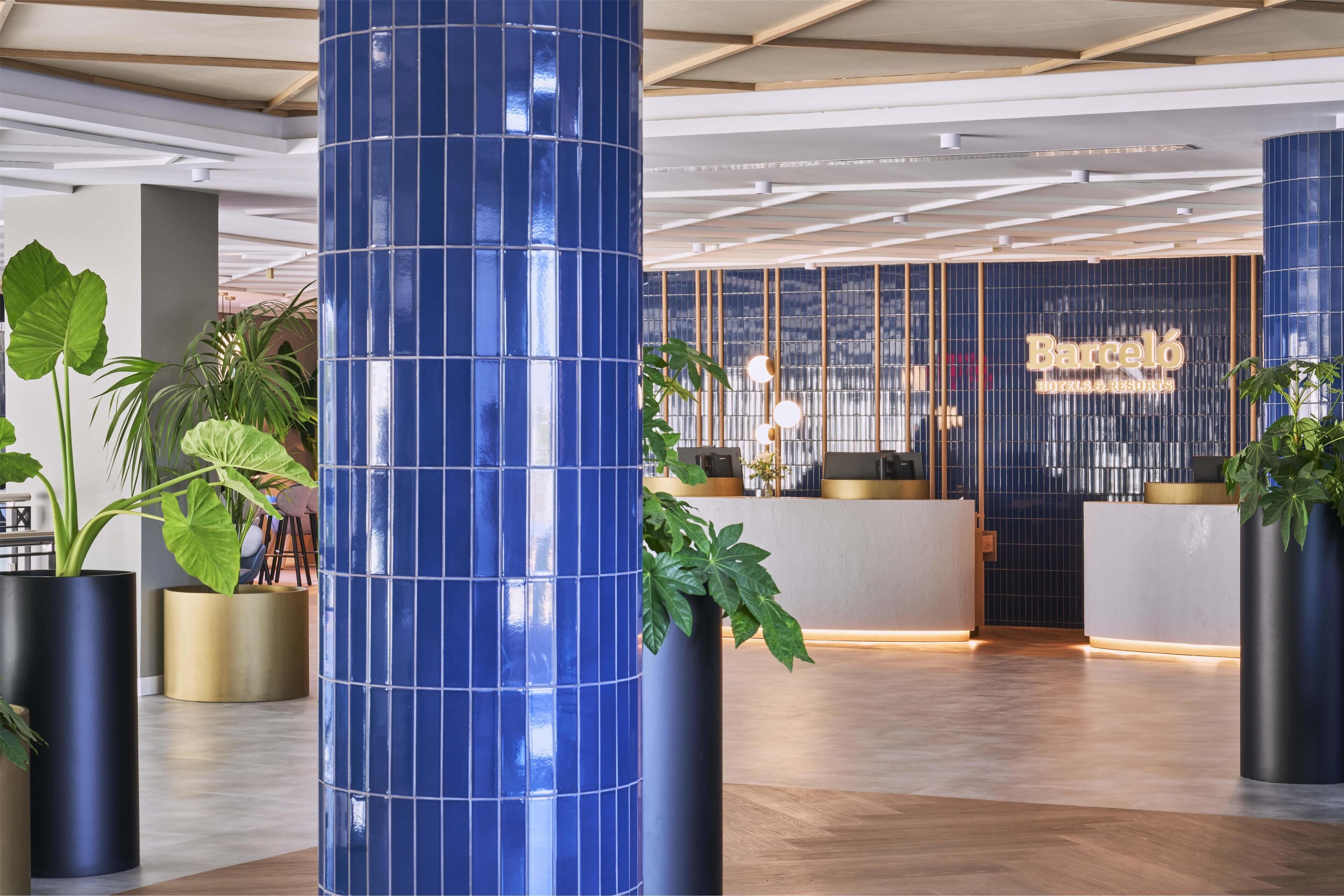 Un moderno vestíbulo de hotel con una recepción, grandes plantas en macetas y un pilar con azulejos azules.