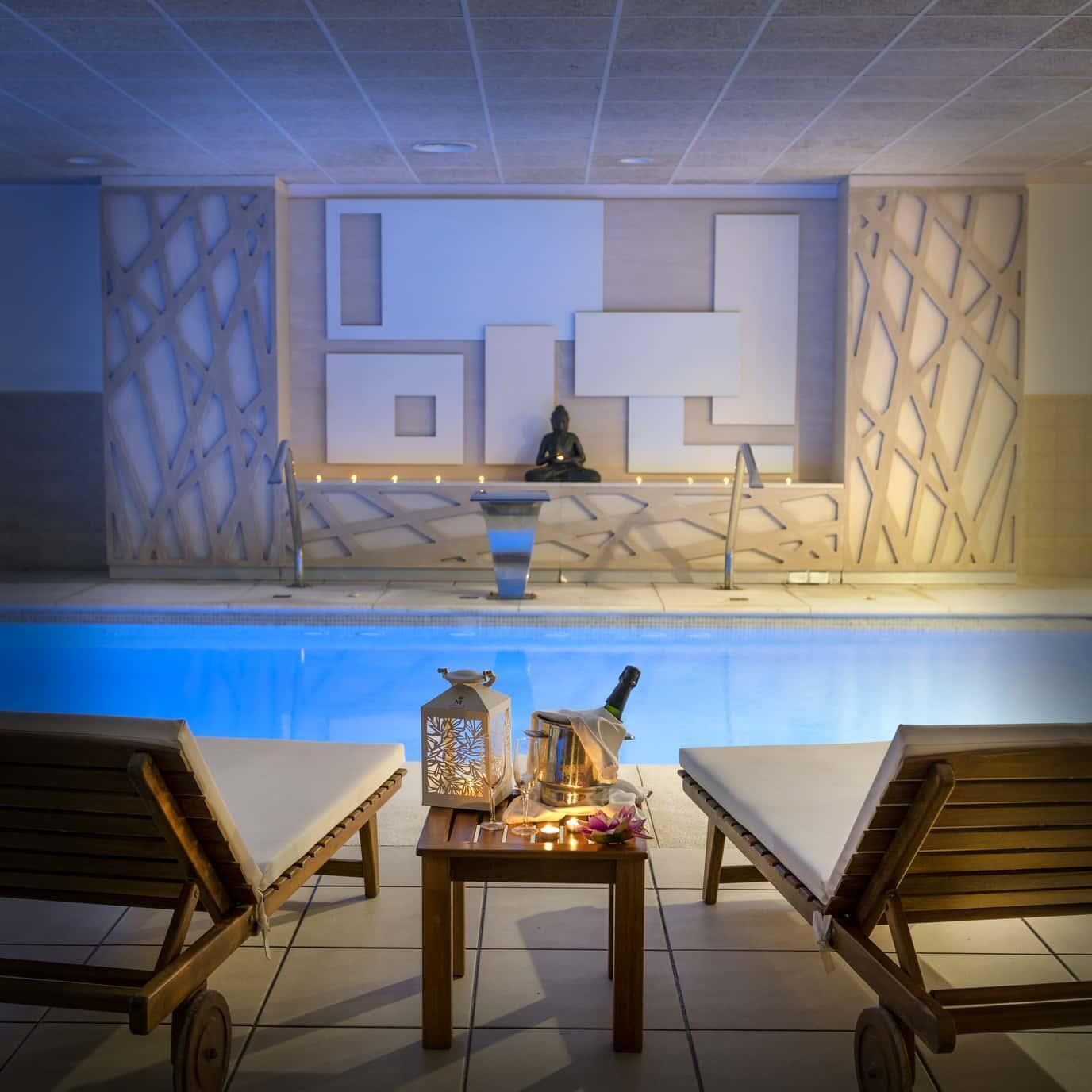 A poolside area with lounge chairs, a table with a bottle and glasses, and a statue in the background.