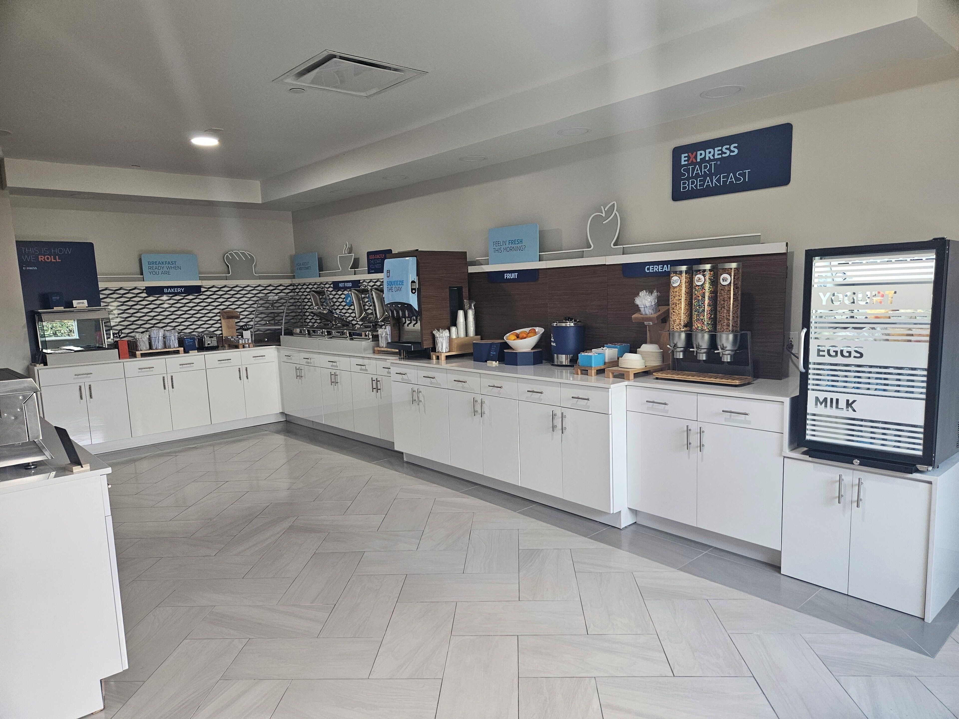 Un moderno comedor para el desayuno con armarios blancos, piso de espiga y diferentes estaciones de desayuno, incluyendo un dispensador de cereales y una máquina para yogur.