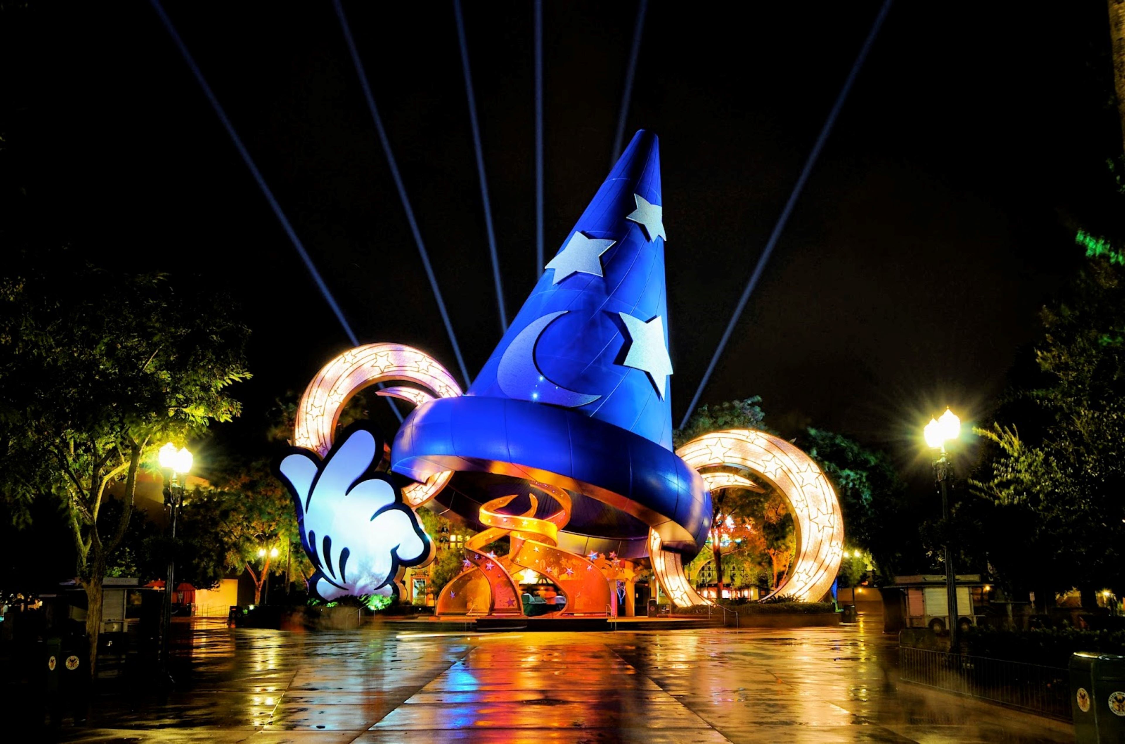 Un'ampia struttura a forma di cappello da mago illuminato, con una falce di luna e stelle, circondata da installazioni luminose circolari.