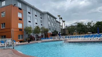 Zona de piscina de hotel con una piscina azul cristalina, sillas de descanso y un edificio de varios pisos con fachada naranja y gris.