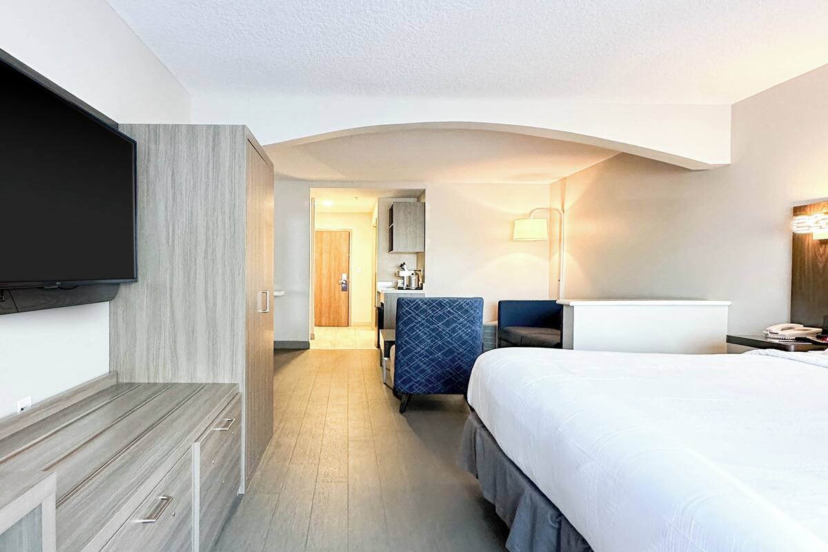 A hotel room with a bed, a TV mounted on the wall, a desk, a chair, and a bathroom visible through an open door.