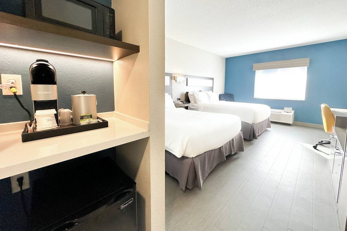 A hotel room with a kitchenette, two beds, a window, and a desk.