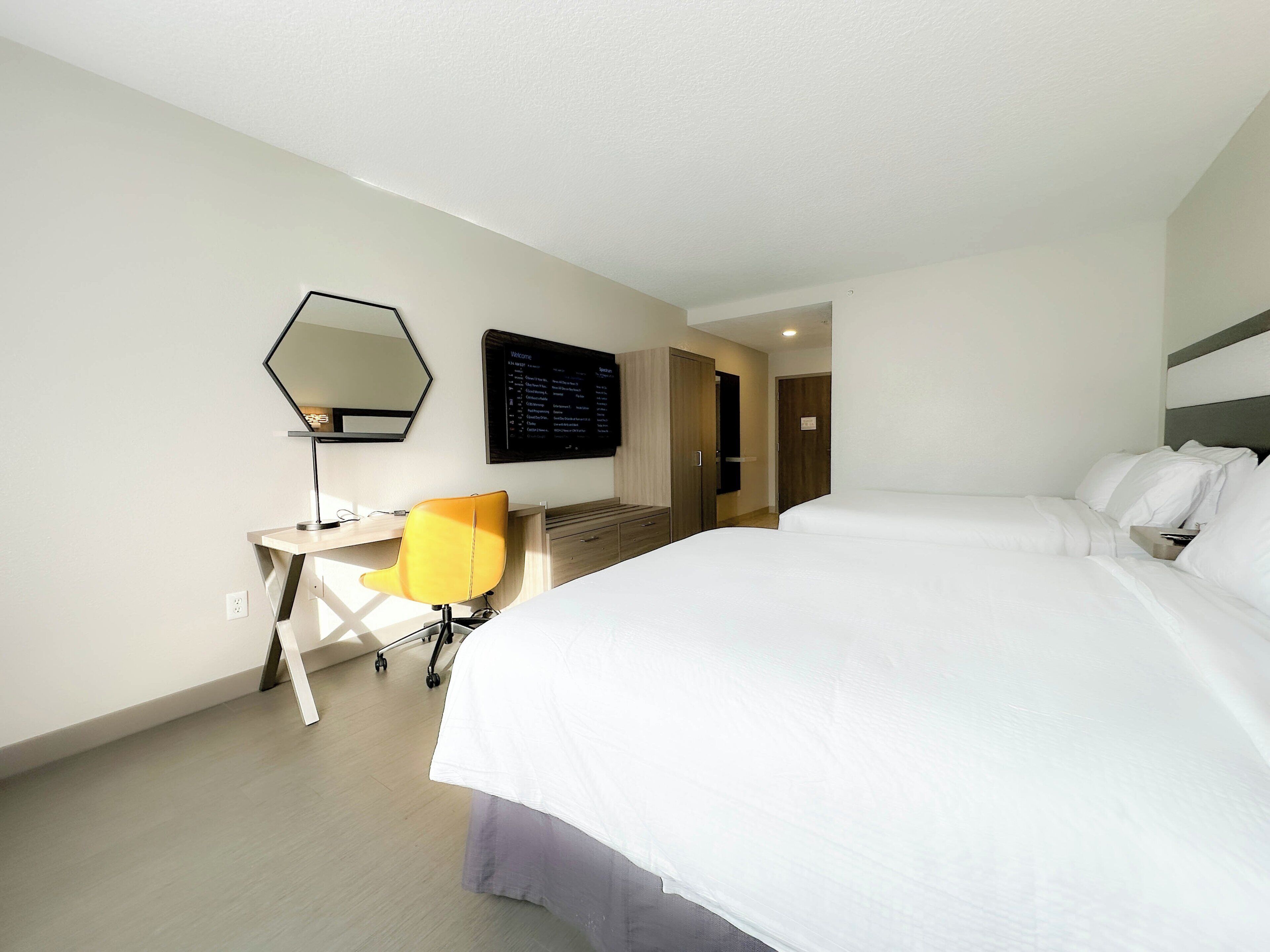 A hotel room with a large bed, a desk with a chair, a mirror, and a digital display on the wall.