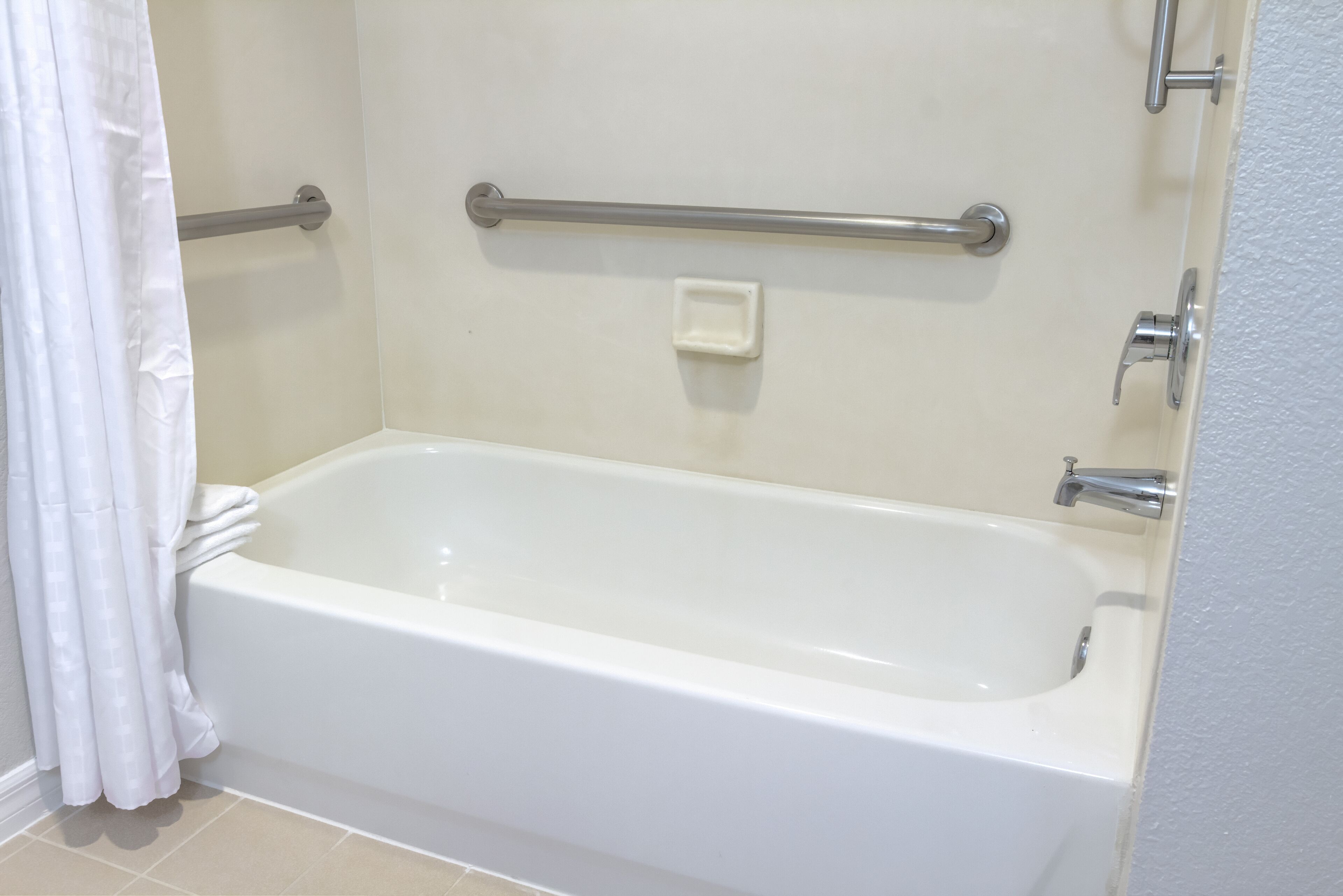 Une baignoire blanche standard, équipée d’une barre d’appui, avec une serviette blanche et un rideau de douche.