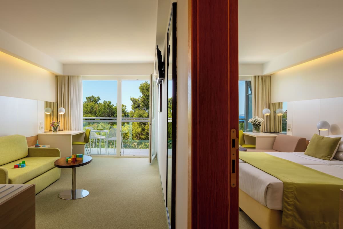 A hotel room with a bed, a sofa, a small table, and a balcony with a view of trees.