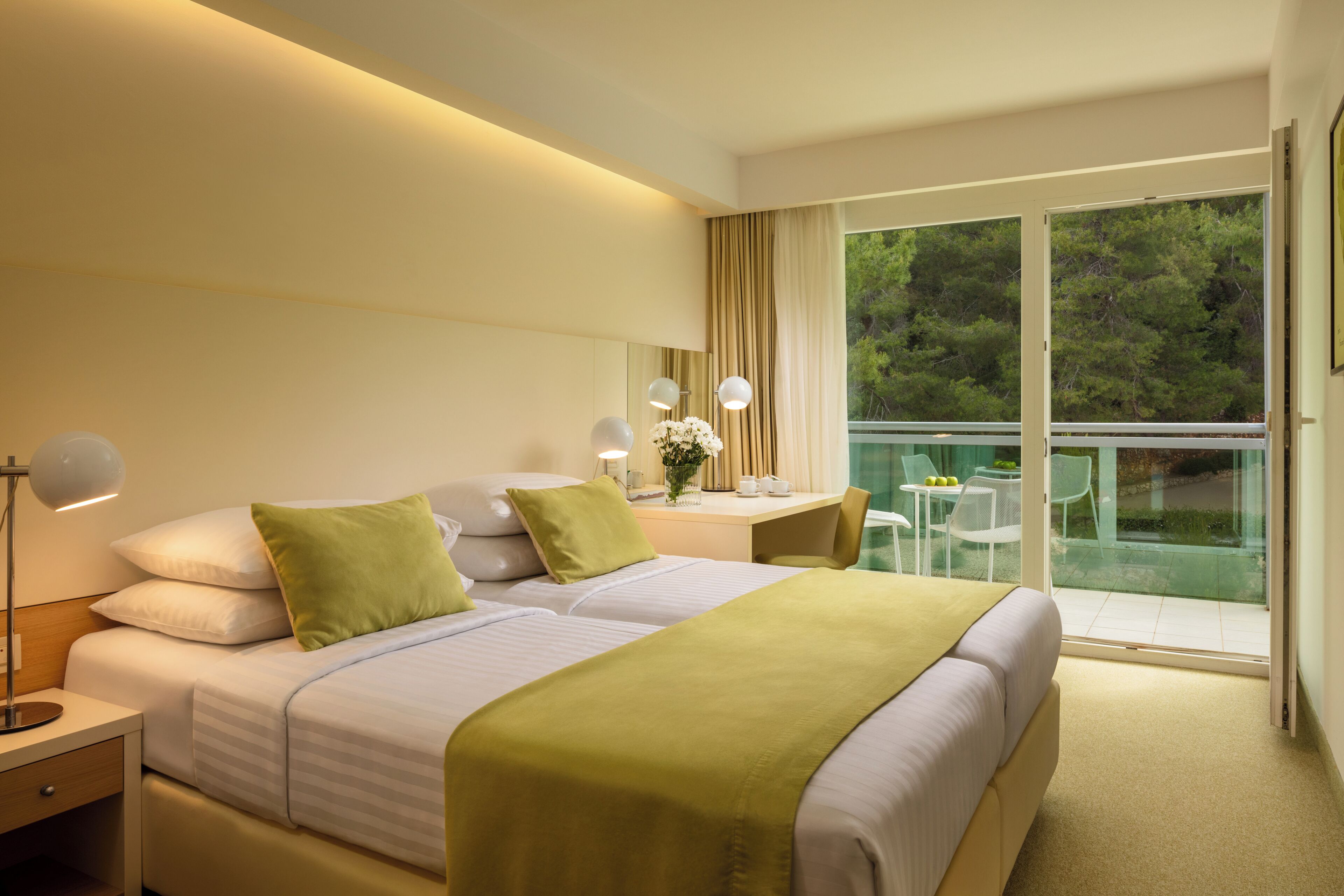 A hotel room with two beds, a desk with a chair, a balcony with glass doors, and a view of trees.