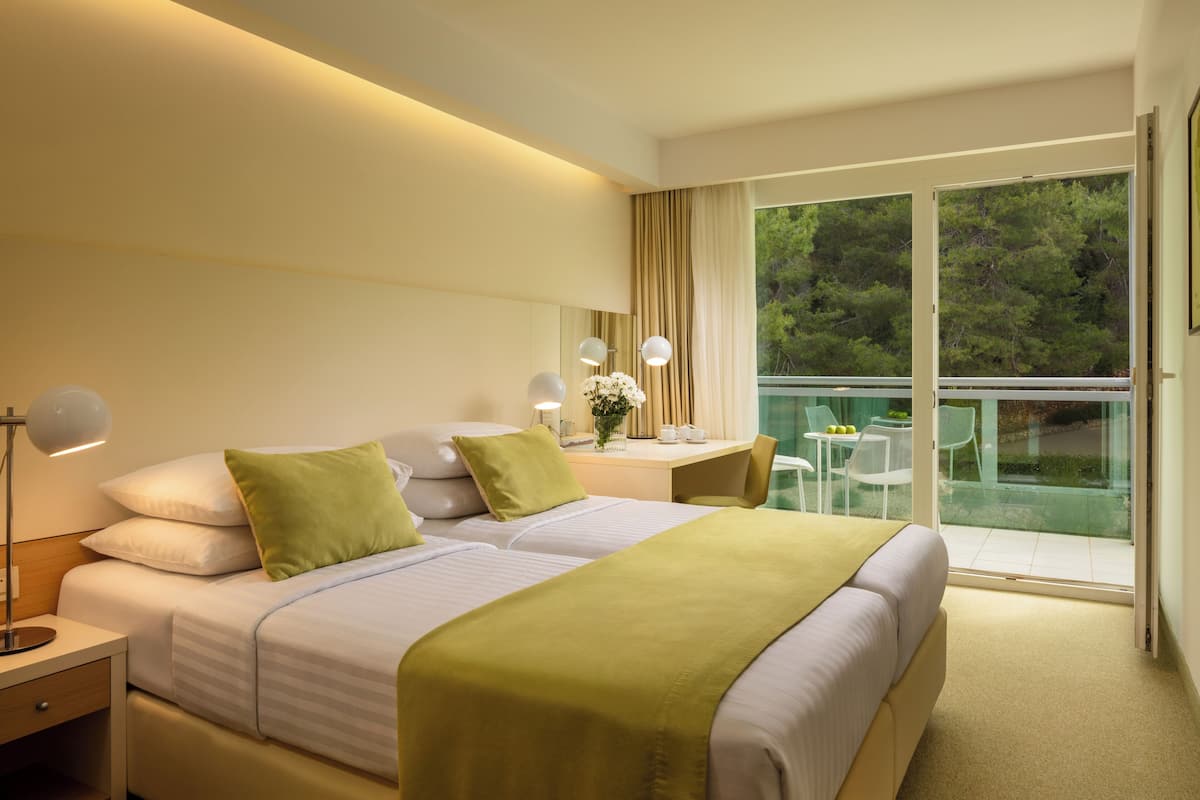 A hotel room with two beds, a desk with a chair, a balcony with glass doors, and a view of trees.