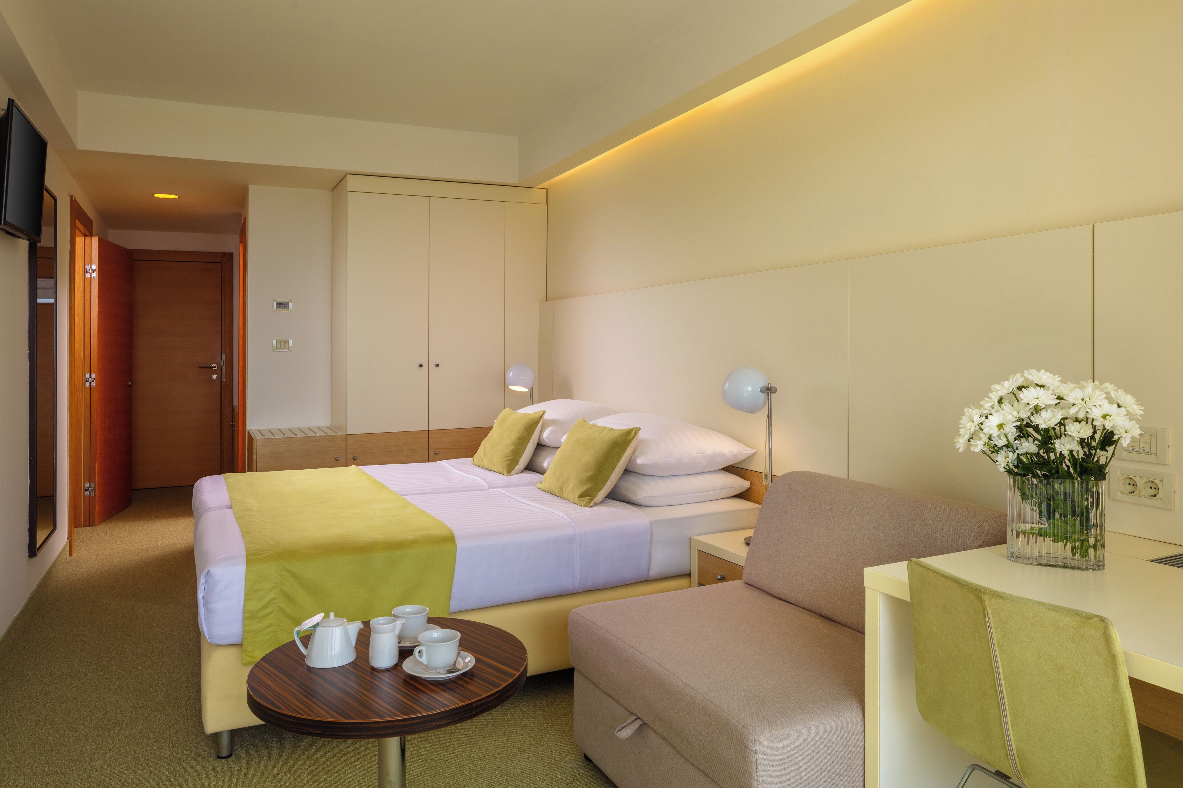 A hotel room with a bed, bedside table, and a small round table with cups and a vase of flowers.