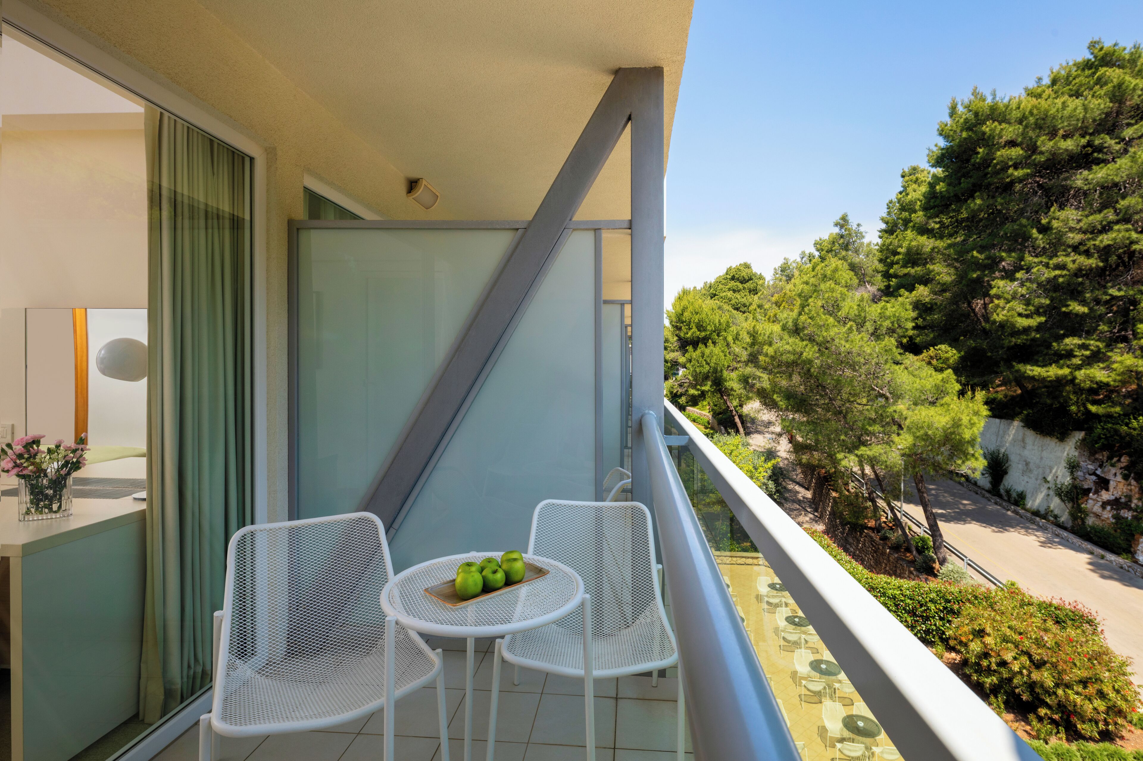 A balcony with a table and chairs, a window with curtains, and a view of trees and a road.