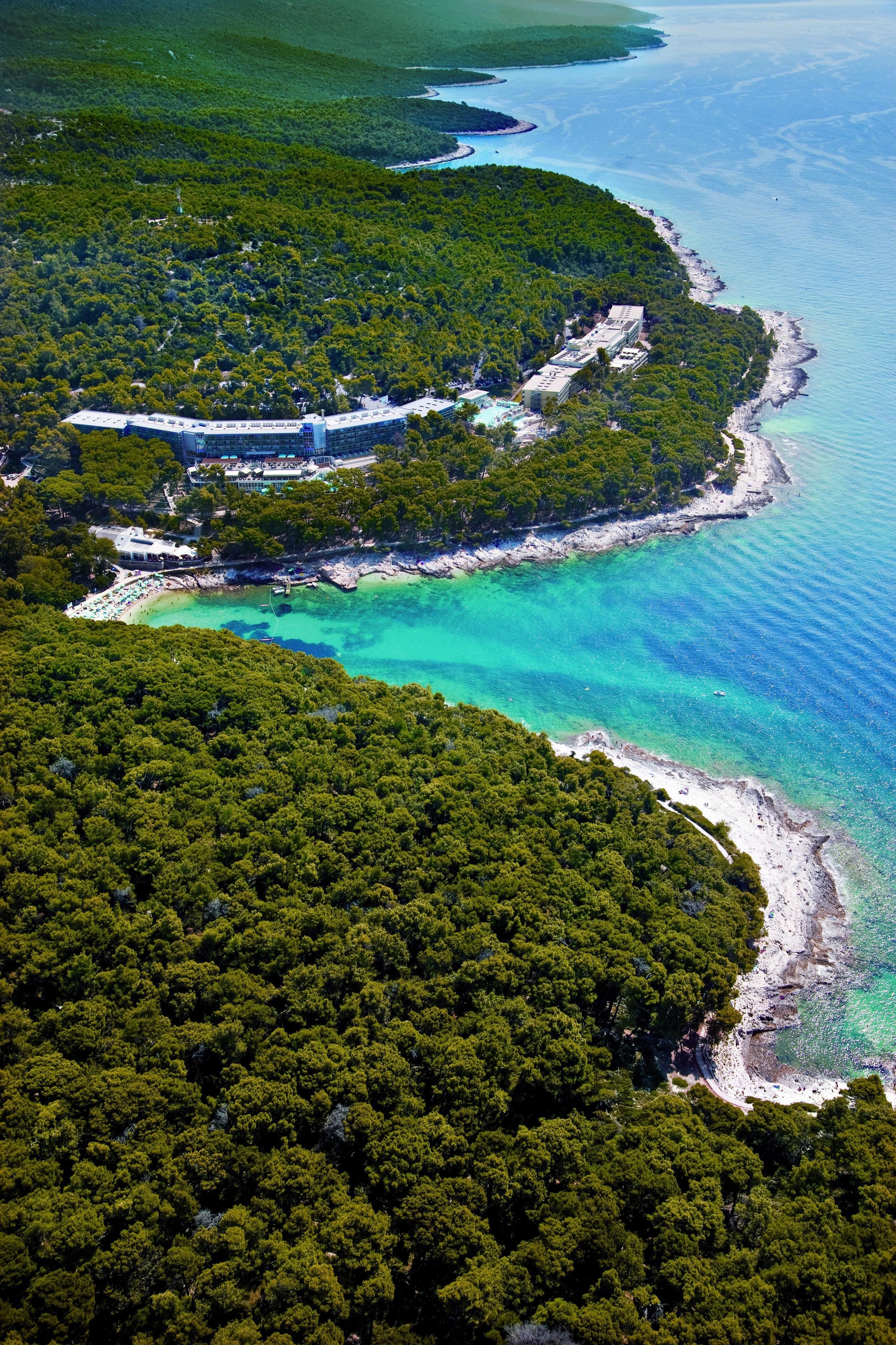 An aerial view of a coastal area with dense forest, a beach, and a large building with multiple levels and a swimming pool.