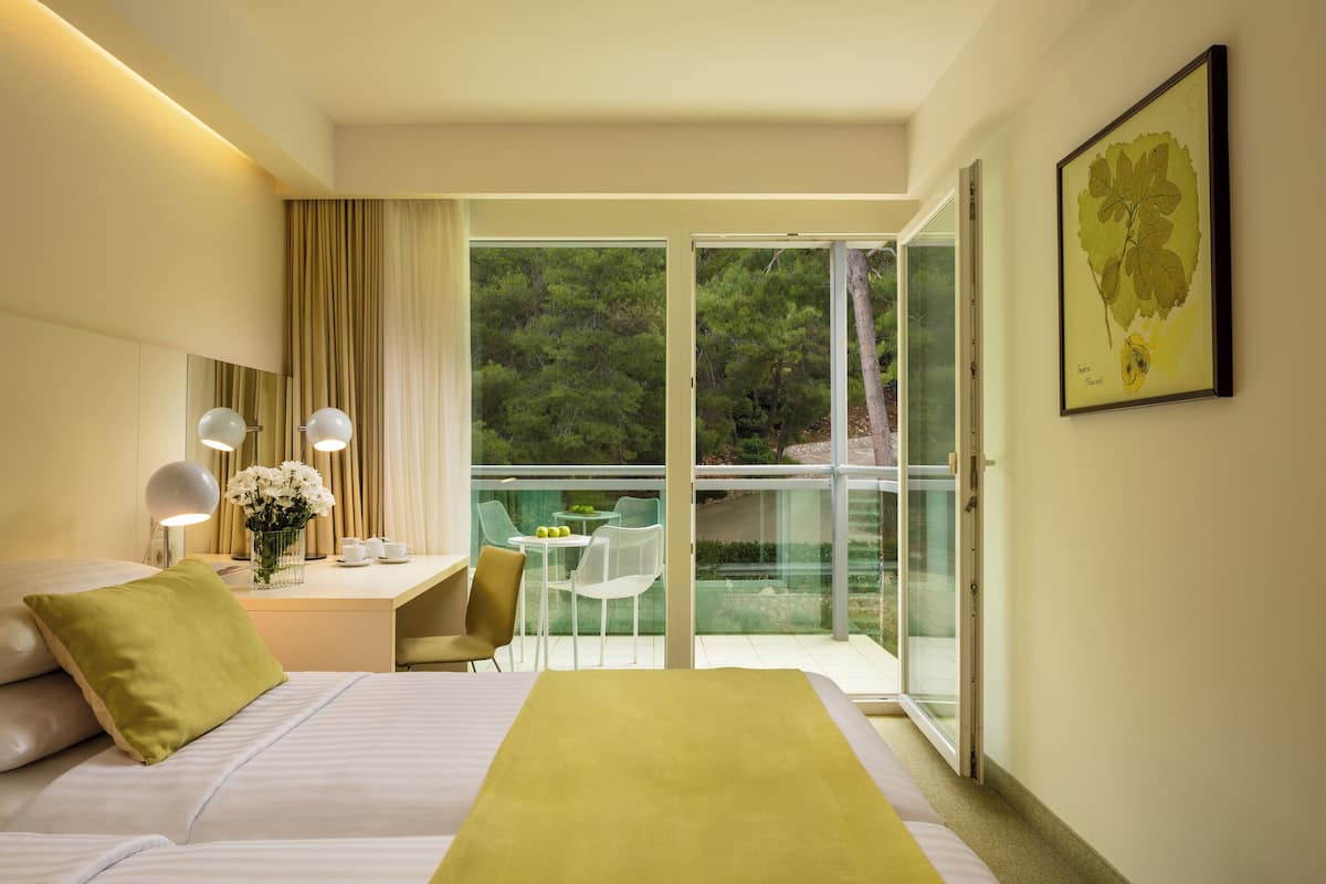 A hotel room with a bed, a desk with a chair, a small table with a vase of flowers, and a balcony with a glass door.