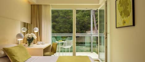 A hotel room with a bed, a desk with a chair, a small table with a vase of flowers, and a balcony with a glass door.